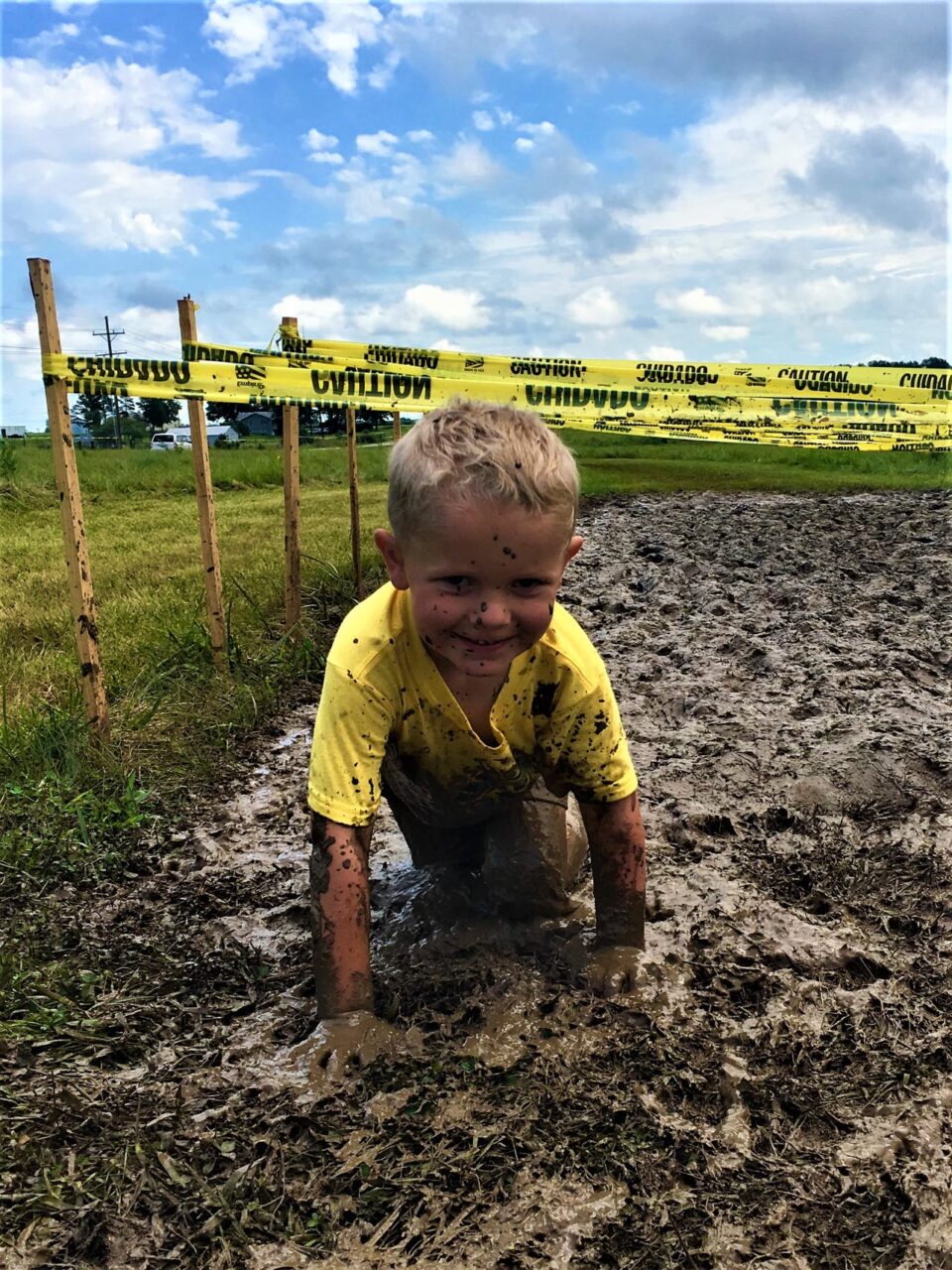 A photograph of a smiling child crawling through the mud, participating in the 'mud mile' at kirksville's family fun weekend