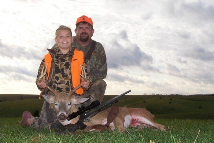A father and daughter pose proudly with the buck they hunted, in a field at dusk