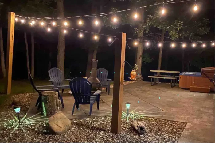 Photograph of the outdoor area of the private farmhouse, at night, showcasing a sitting area with patio furniture, a hot tub, and string lighting overhead
