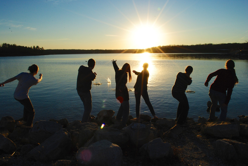 A photograph of six people standing on a shoreline skipping rocks at sunset